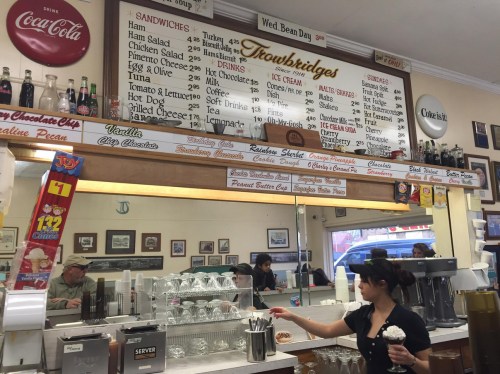 A diner counter with a menu board, staff serving ice cream, and customers enjoying their meals in a nostalgic setting.