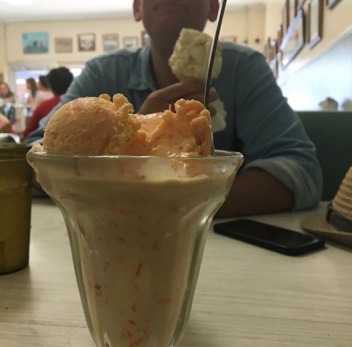 A glass of orange ice cream with a spoon, with a person in the background holding a cone.
