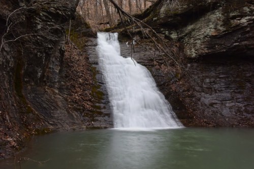 A serene waterfall cascading over rocky cliffs into a calm, green pool surrounded by trees and autumn leaves.