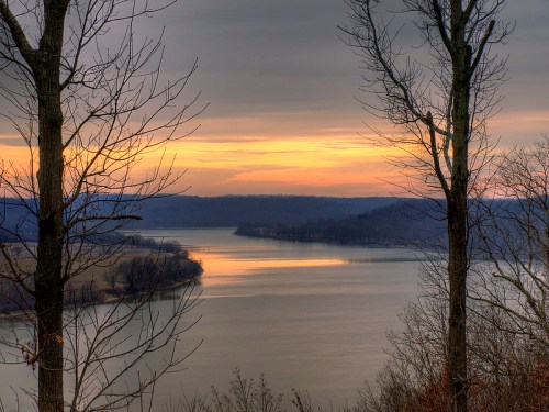 A serene river landscape at sunset, framed by bare trees and soft clouds reflecting on the water's surface.