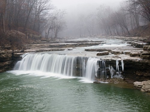 A serene waterfall cascades over rocky ledges into a misty, green river surrounded by bare trees.