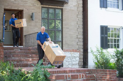 Two delivery workers carrying large boxes down the steps of a house. One uses a dolly for easier transport.