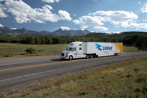 A white semi-truck with a blue and yellow logo drives along a highway surrounded by mountains and greenery.