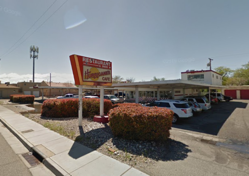 Sign for Hurricane's Cafe in front of a parking lot with several cars and a building in the background. Clear blue sky.