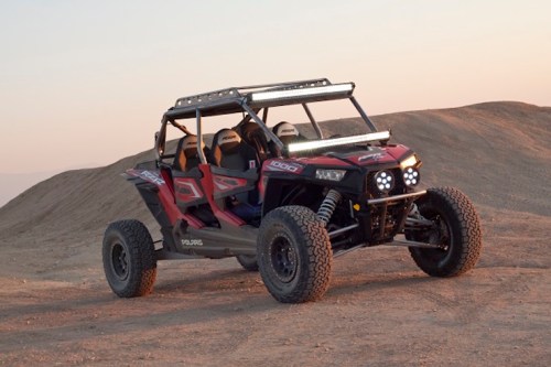 A red off-road vehicle parked on a sandy terrain during sunset, featuring a light bar and rugged tires.