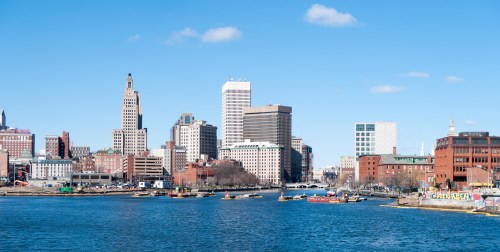City skyline with tall buildings along a river, blue sky, and boats on the water.