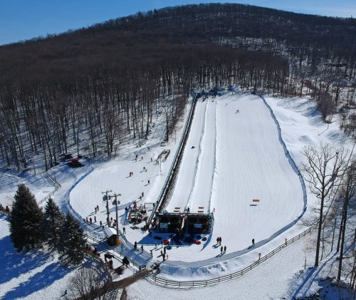 Aerial view of a snowy ski slope with people skiing and a ski lift in a wooded area on a sunny day.