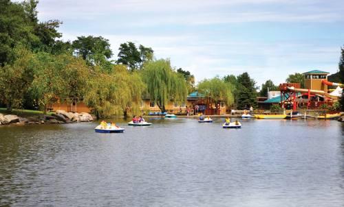 People paddle boats on a calm lake surrounded by trees and colorful buildings in the background.