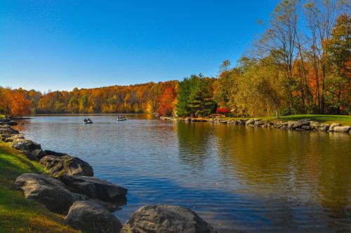 A serene lake surrounded by colorful autumn trees, with two people kayaking in the distance under a clear blue sky.