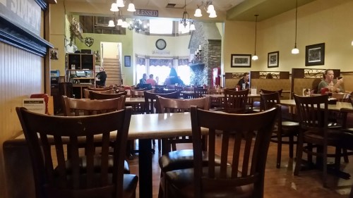 A cozy restaurant interior with empty tables and chairs, featuring a staircase and patrons in the background.