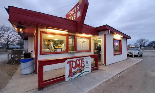 A small drive-in restaurant with a red roof and sign, featuring a menu board and a car parked outside.