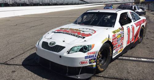A white NASCAR race car with the number 11 parked on a racetrack, surrounded by empty grandstands.