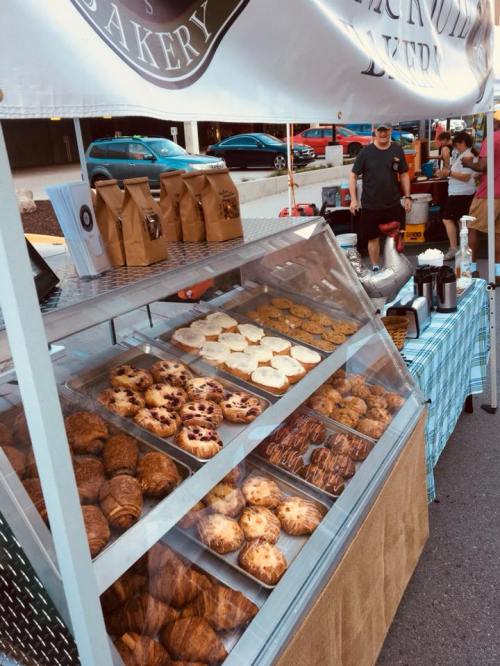 A bakery stand at a market displaying various pastries and baked goods, with customers in the background.