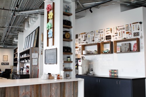 A modern café interior featuring a wooden counter, shelves with vintage kitchen items, and a minimalist design.