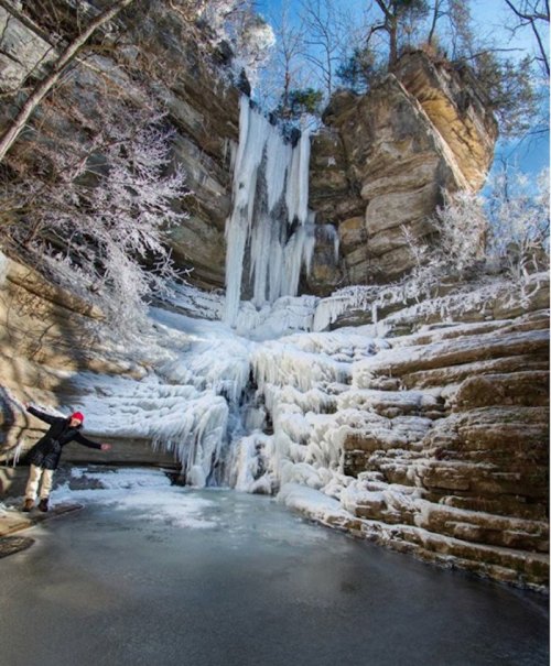 A frozen waterfall surrounded by icy cliffs and trees, with a person standing on the ice below.