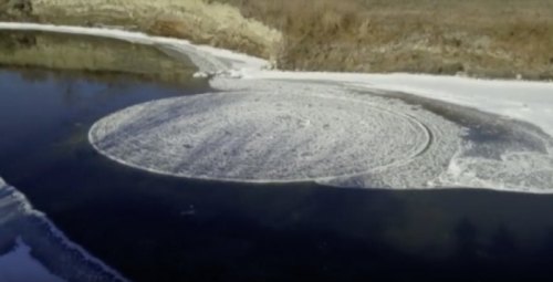 A circular pattern of ice and snow forms on the surface of a river, surrounded by bare land and trees.