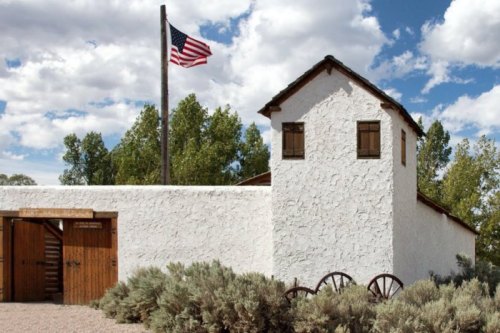 A white adobe building with a wooden tower, an American flag, and surrounding greenery under a partly cloudy sky.