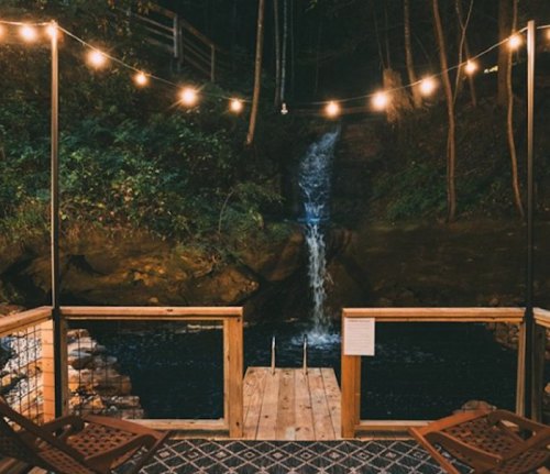 A serene wooden deck with string lights overlooking a small waterfall and dark water below, surrounded by trees.