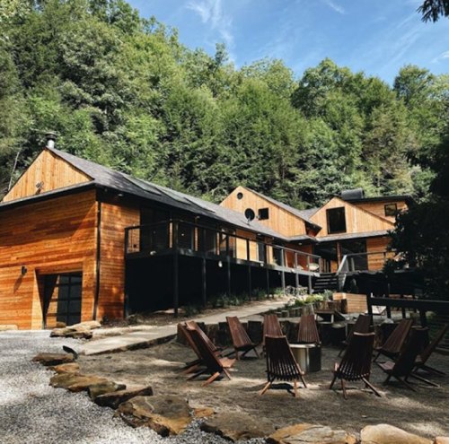 A modern wooden cabin surrounded by trees, featuring a spacious deck and outdoor seating area with chairs.