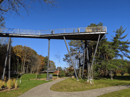 A walkway elevated on stilts above a park, with people walking and trees in the background under a clear blue sky.