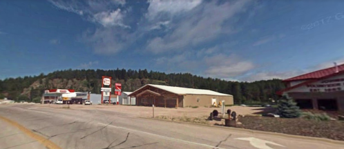 A small town scene featuring a gas station and shops along a road, with trees and hills in the background under a blue sky.