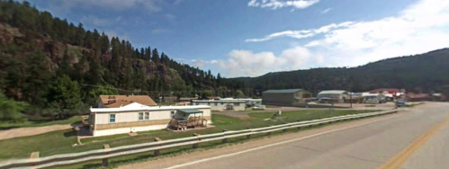 A small town scene with houses, trees, and mountains under a clear blue sky. A road runs alongside the area.