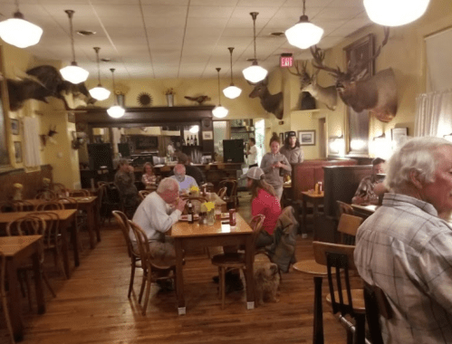 A cozy restaurant interior with wooden tables, patrons dining, and mounted animal heads on the walls. Warm lighting.