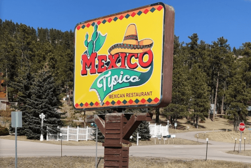 Colorful sign for "Mexico Tipico," a Mexican restaurant, surrounded by trees and a clear blue sky.