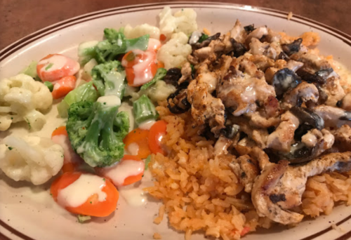 A plate of seasoned chicken over rice, accompanied by steamed broccoli, cauliflower, and carrots drizzled with dressing.