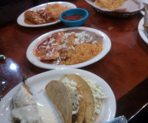 A table with plates of Mexican food, including tacos, enchiladas, rice, and beans, alongside a bowl of salsa and chips.