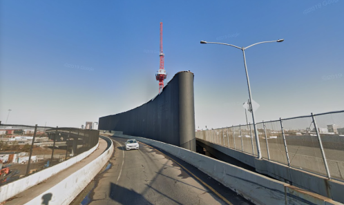 A highway curve with a tall black wall and a red tower in the background under a clear blue sky.