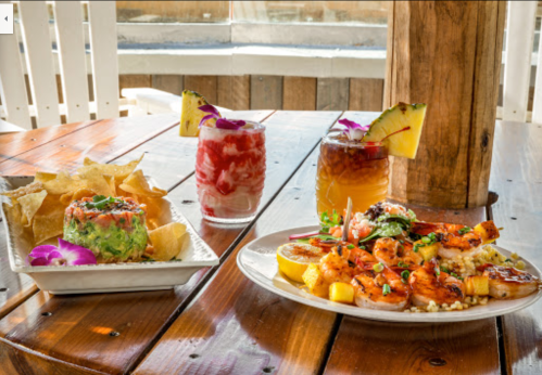 A table with shrimp dish, guacamole, tortilla chips, and two colorful cocktails garnished with fruit and flowers.