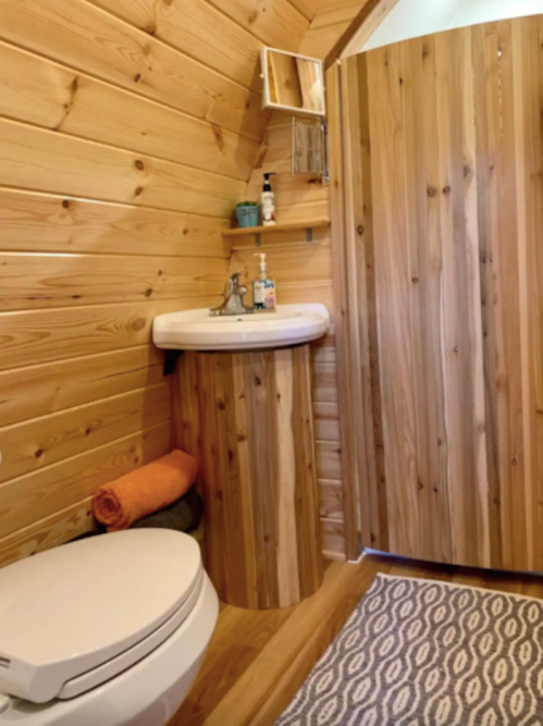 Cozy wooden bathroom featuring a round sink, toilet, and a shower with wooden paneling. Soft towels and a rug add warmth.