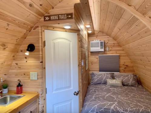 Cozy wooden interior of a small cabin with a bed, kitchenette, and a sign for hiking trails.