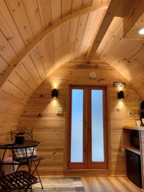 Cozy wooden interior of a small cabin with arched ceiling, a table, and a door leading outside. Warm lighting enhances the ambiance.