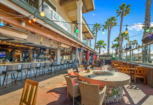 Outdoor bar and dining area with colorful lights, palm trees, and a fire pit, set against a sunny sky.