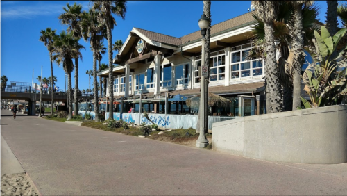 A beachfront restaurant with large windows, surrounded by palm trees and a paved walkway along the shore.