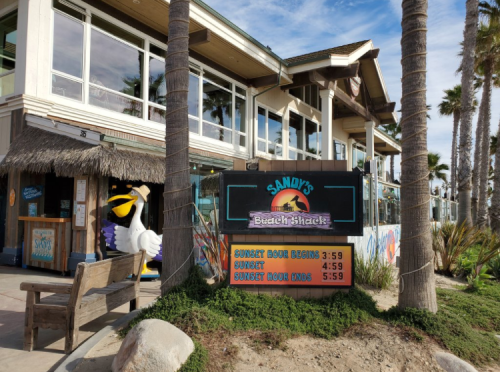 A beachside restaurant sign displays sunset hours and a colorful exterior with palm trees in the background.