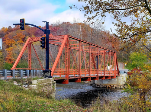 A red metal bridge spans a river, surrounded by autumn foliage and a traffic light nearby.