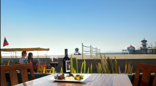 A beachside dining scene with a table set for two, wine, and a view of the pier and ocean under a clear blue sky.
