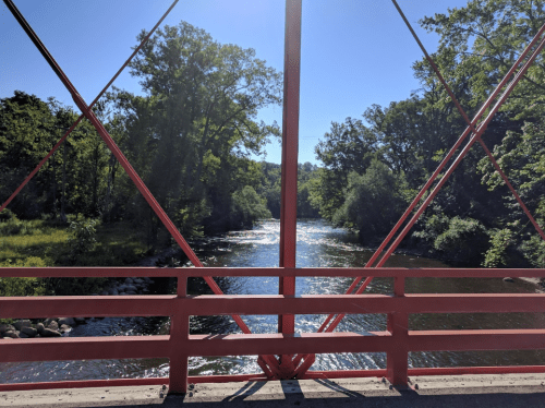 A view of a river through a red bridge's railing, surrounded by lush green trees under a clear blue sky.