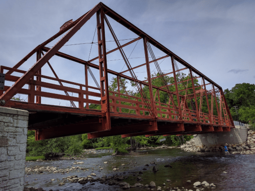 A red metal bridge spans a river, surrounded by trees and rocky banks under a cloudy sky.
