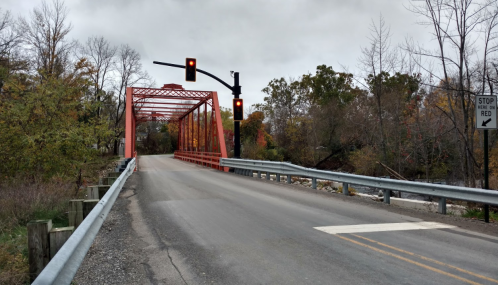 Red metal bridge with traffic lights, surrounded by trees in autumn colors, under a cloudy sky.
