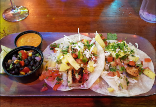 Two tacos filled with grilled chicken, pineapple, and toppings, served with black beans and a side of sauce.