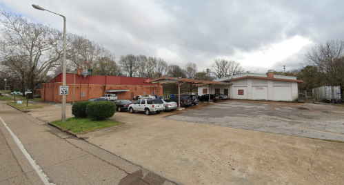 A parking lot with several cars outside a red brick building under a cloudy sky. Trees line the background.