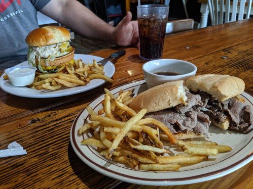A table with a plate of fries and a sandwich filled with beef, alongside a burger and more fries in the background.