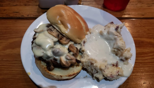A plate with a burger topped with mushrooms and creamy sauce, alongside a serving of mashed potatoes with gravy.
