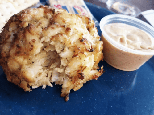 A golden-brown crab cake sits on a blue plate next to a small cup of creamy dipping sauce.