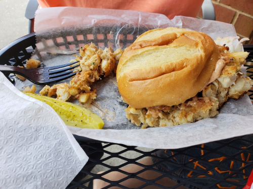 A crab cake sandwich on a bun with a side of pickles, served on a paper-lined basket.