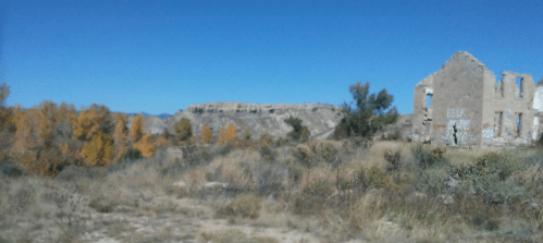 A desolate landscape featuring a crumbling building, colorful autumn trees, and distant rocky hills under a clear blue sky.